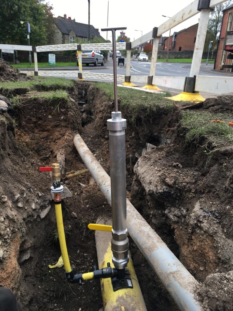 Construction site with exposed earth and pipes in a trench. A metal tool is inserted into a yellow pipe, surrounded by temporary barriers. Urban setting.