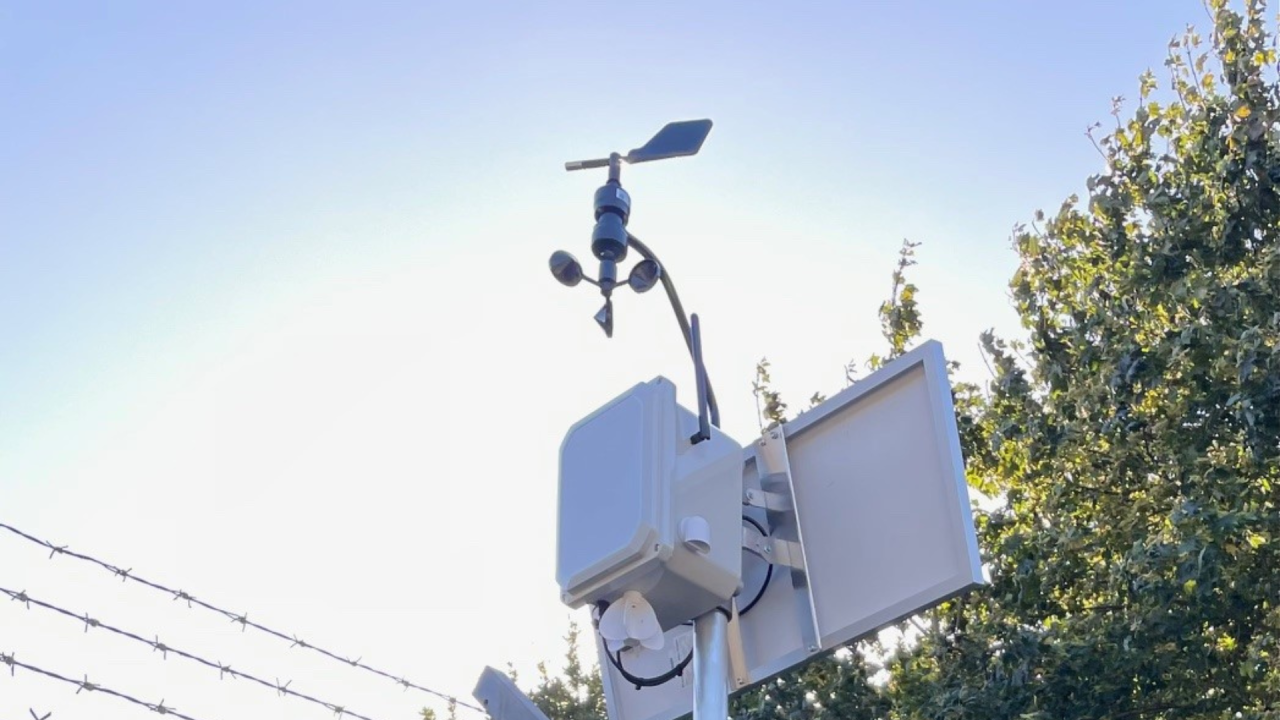 A fixed emissions monitoring device equipped with a solar panel and weather instrumentation mounted on a pole near trees and security fencing, used for continuous gas detection.