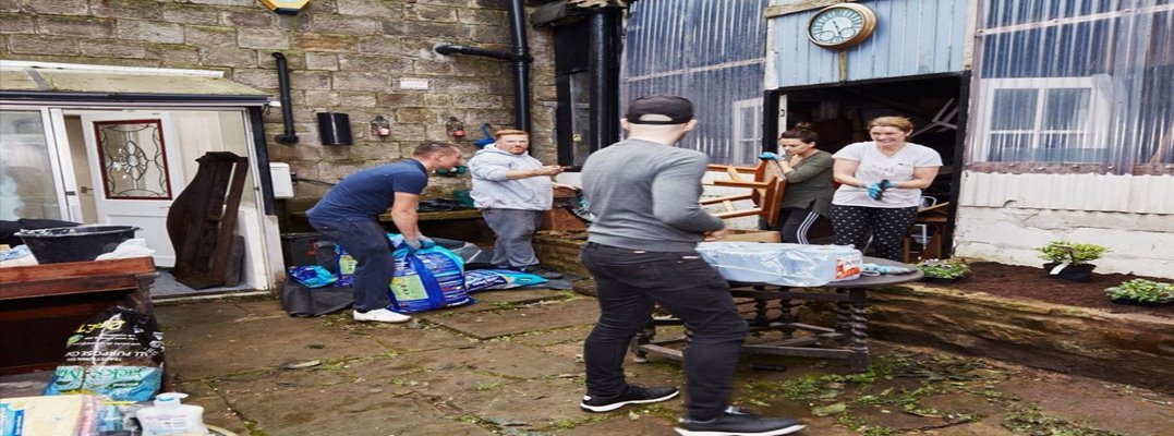 People working together in an outdoor area, moving bags of soil and furniture near a rustic building with a clock above the doorway and plants along the wall.