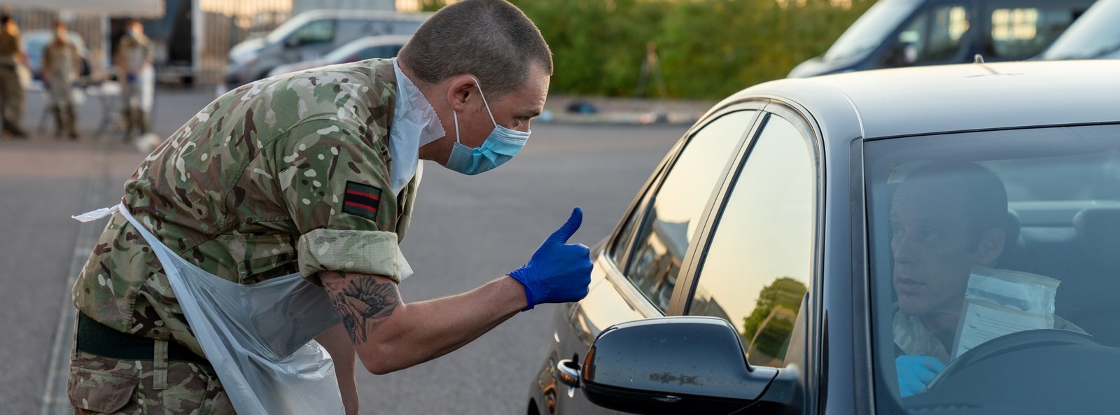 A soldier in camouflage, wearing a mask and gloves, leans towards a car window at a drive-through test site, giving a thumbs-up to the driver.