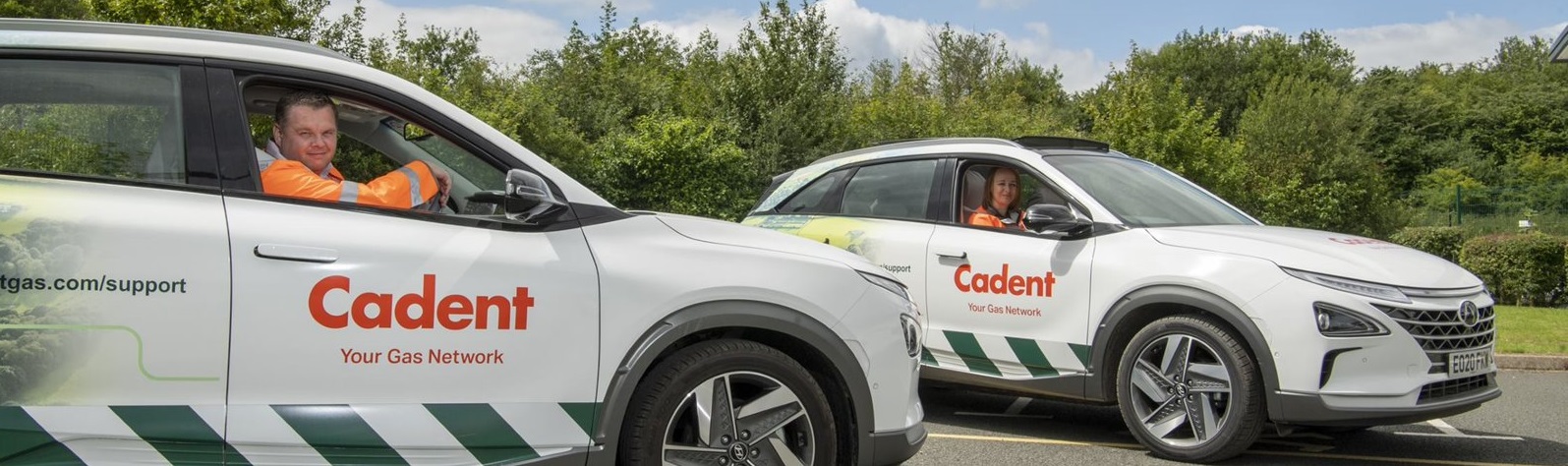 Two Cadent-branded vehicles parked on a road, with drivers in orange uniforms smiling. Background of lush greenery under a partly cloudy sky.