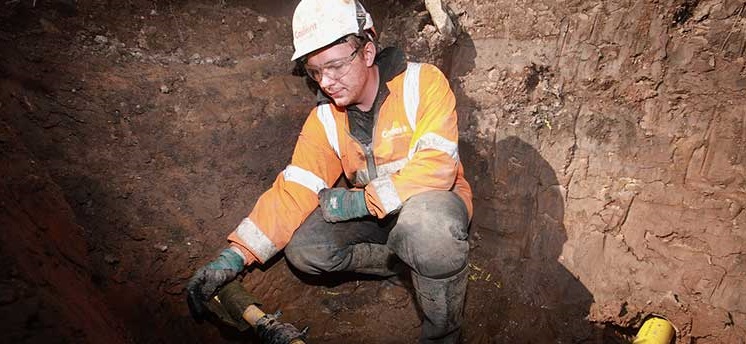 A construction worker in an orange safety jacket and white helmet kneels in a trench, inspecting yellow pipes. The setting appears to be an outdoor excavation site.