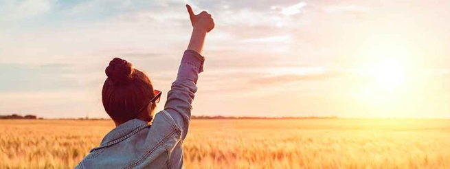 A woman stands in a sunlit field of tall grass, facing a bright sunset. She wears a denim jacket and gives a thumbs-up, evoking a sense of positivity and freedom.