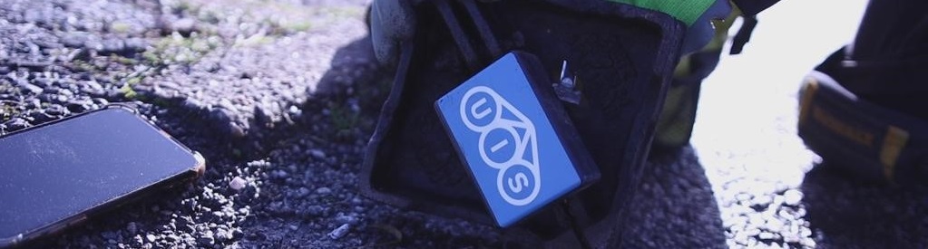 A person wearing green gloves installs a blue sensor in a small hole on a textured road surface. A smartphone lies nearby. The scene is lit with a cool, soft light.