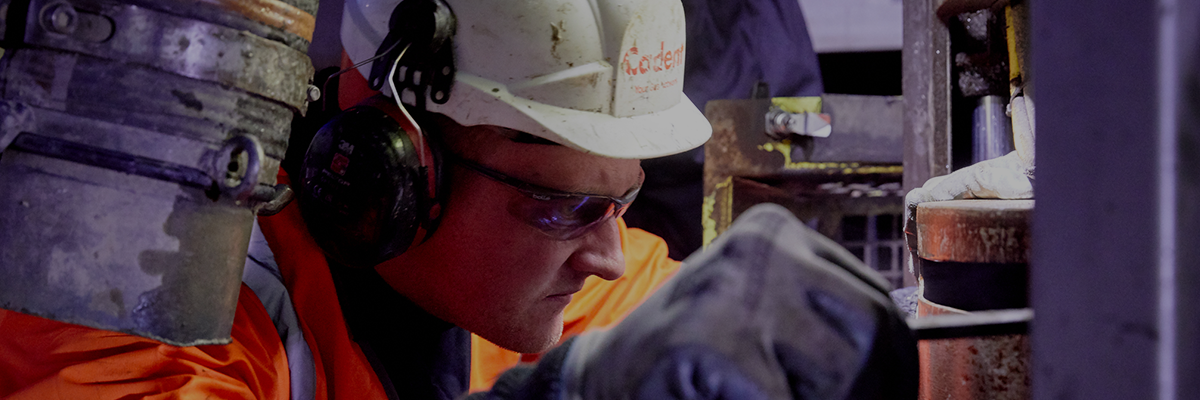 A focused worker in a hard hat and ear protection operates machinery, wearing orange safety gear. The scene conveys concentration and professionalism.