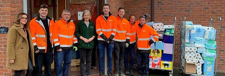 A group of eight people, some in bright orange jackets, stand smiling by a brick building entrance. Beside them are carts filled with various household supplies like toilet paper and detergent, suggesting a donation or community event.