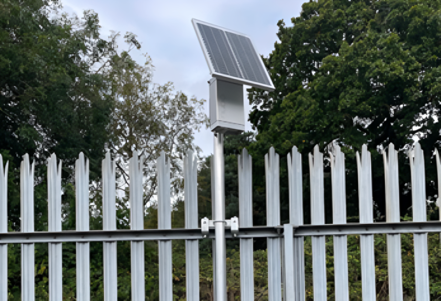 A small solar panel mounted on top of a metal box fixed to a tall metal fence, surrounded by trees in the background.