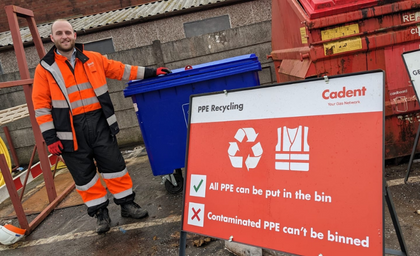 A person in an orange and black work outfit stands next to a blue recycling bin. A sign reads,