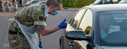 A masked military personnel in camouflage and gloves gives a thumbs-up to a person in a car at a drive-through testing site. Tents and vehicles are in the background.