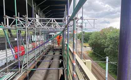 Scaffolding on a bridge under construction, with a worker in orange safety gear. Overcast sky, rail tracks, and trees beside the bridge.