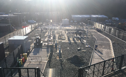 Industrial site with fenced areas, equipment, and gravel ground under bright sunlight, with buildings and vehicles in the background.