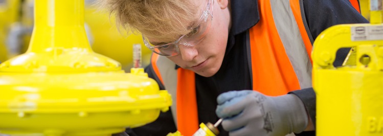 A focused worker in safety gear inspects machinery. Wearing an orange vest and goggles, the person examines a yellow industrial component with precision.