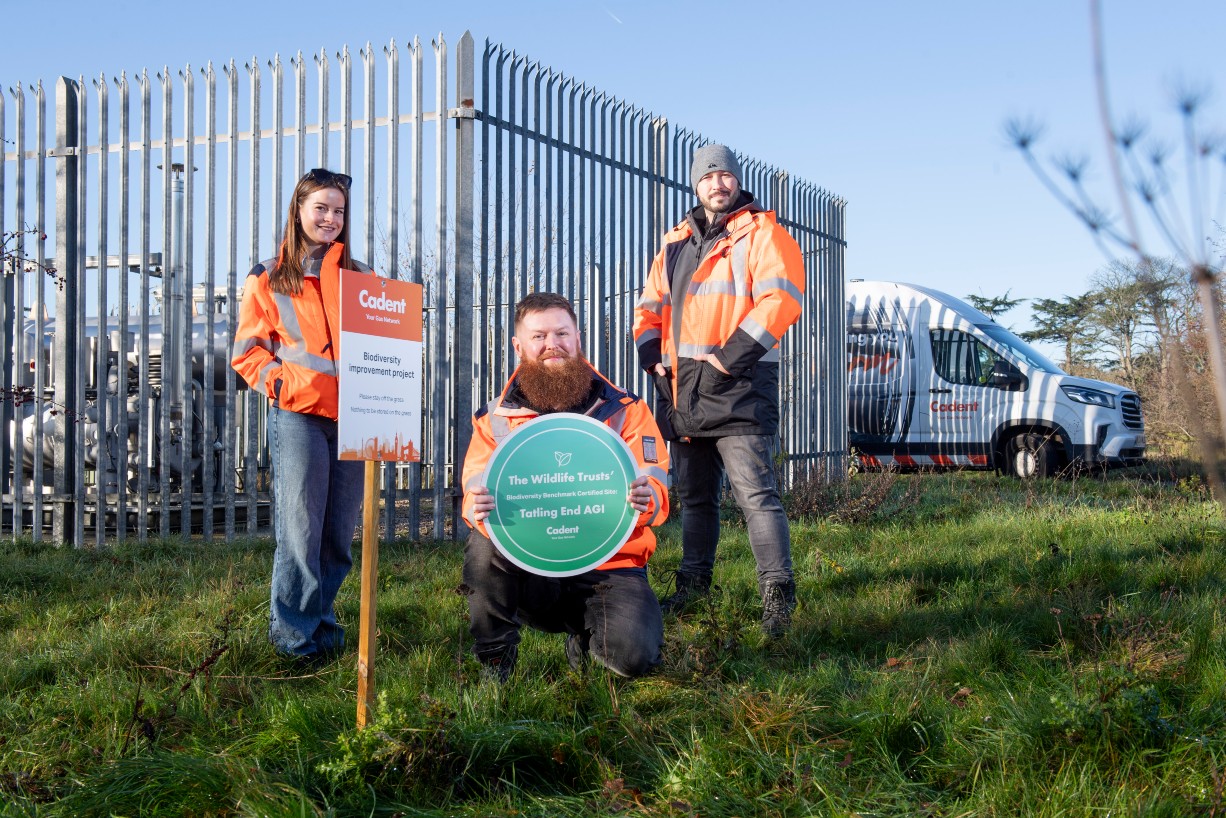Three Cadent workers wearing high-visibility orange jackets stand and kneel on grass beside a metal security fence at a gas site.