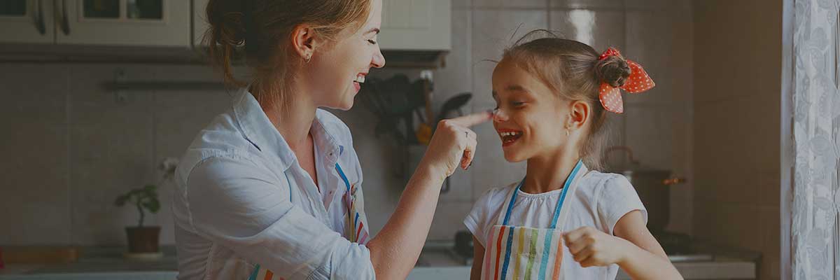 A woman and a girl are interacting in a bright kitchen, both wearing colorful aprons. The woman is poking the girl's nose playfully