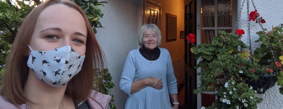A woman wearing a penguin-printed mask takes a selfie with an elderly woman standing in a doorway. They are surrounded by plants and flowers, conveying warmth and connection.