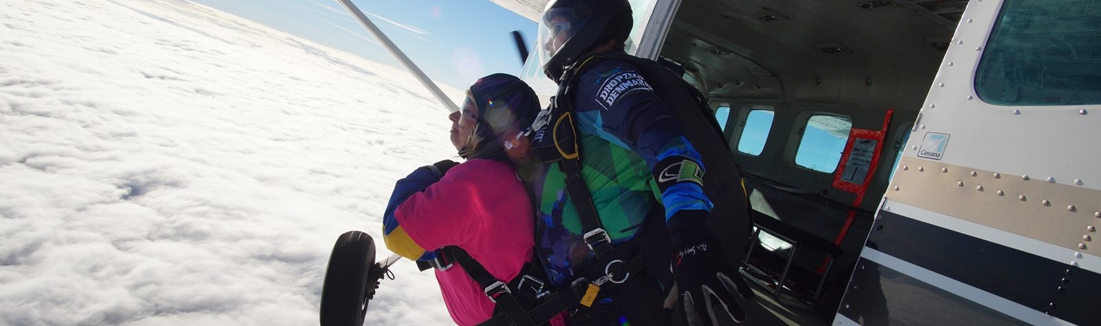 A tandem skydiver pair leans out of an open airplane door, poised to jump. The skydivers wear colorful suits against a backdrop of a cloudy sky.