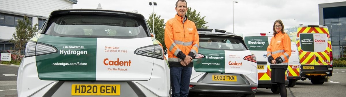 Two people in orange jackets stand beside Cadent-branded hydrogen cars in a parking lot. A Cadent utility van is in the background. The mood is professional.