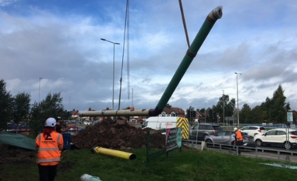 Construction workers in orange vests oversee a large green pipe being lifted by a crane near a busy road, with cars and cloudy skies in the background.