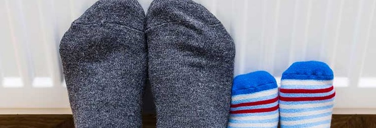 Two pairs of feet in cozy socks, one gray and argyle, the other blue with stripes, rest on a warm radiator. The scene conveys warmth and comfort.