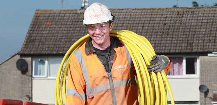 A construction worker, wearing an orange safety jacket and helmet, smiles while carrying a coiled yellow cable. Red barriers and a house are in the background.