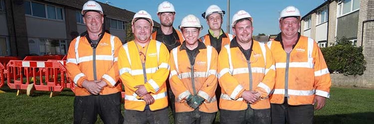 Seven construction workers stand on a grassy area in front of houses, wearing orange safety jackets and white helmets, smiling in the sunlight_banner_image