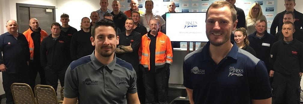 Two men in branded shirts stand in front of a group of people in a conference room. A screen displays a logo. The mood is professional and upbeat_Banner_image