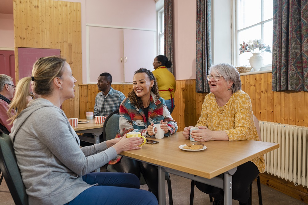 Three women sit at a table in a cozy room, smiling and chatting over mugs of tea. A plate with biscuits is on the table. A warm, friendly atmosphere.