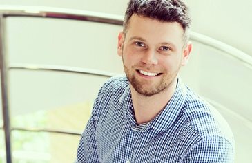 A smiling man with short, dark hair and a beard wears a blue checkered shirt. He is sitting on a staircase with metal railings, conveying a cheerful tone.