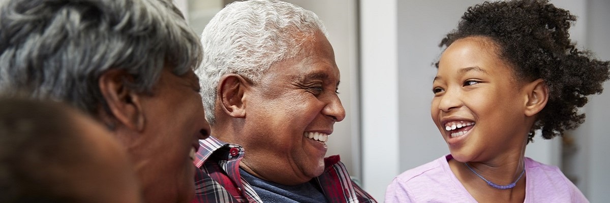 Three people sitting close, smiling warmly at each other. An older man with short white hair and a young girl with curly hair exchange happy expressions. Another person with grey hair is partially visible on the left. The setting seems casual and joyful.