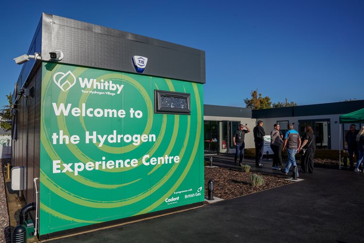 Green building sign reads 'Welcome to the Hydrogen Experience Centre' under blue sky. People gather outside, suggesting a community event.