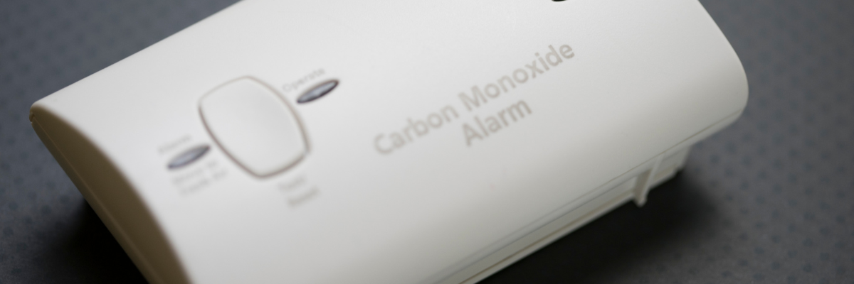 Close-up of a carbon monoxide alarm with a white casing, featuring a test/reset button and indicator lights. The label Carbon Monoxide Alarm is printed on the device. The background is a dark, textured surface.