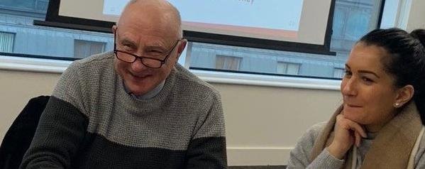 Two people sat at a table having a conversation. An older gentleman with a woman in her mid-twenties smiling.