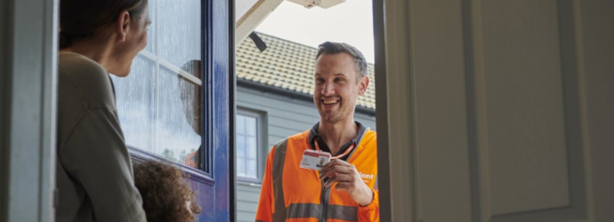 A Cadent engineer in an orange hi-vis vest stands at a doorway holding an ID while speaking to a resident inside. 