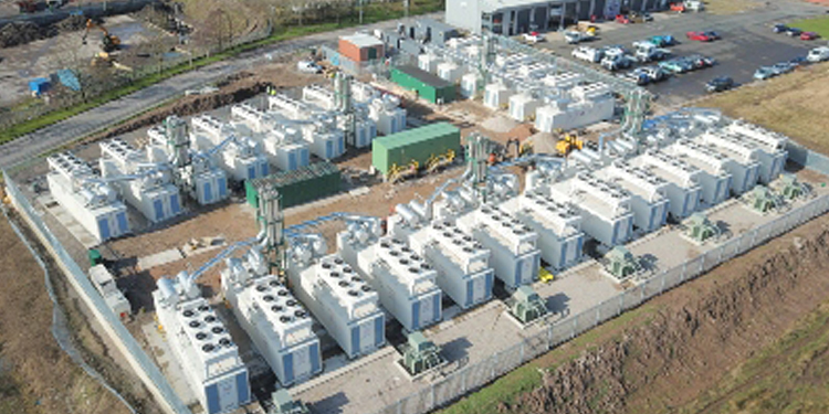 Aerial view of an industrial site with rows of large white units, pipes, and ventilation systems, surrounded by fencing and adjacent to a parking area.