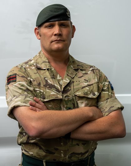 Serious soldier in camo uniform and green beret crosses arms, standing confidently against a plain backdrop, conveying strength and authority.