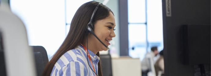 A person wearing a headset is smiling while looking at a computer screen in an office setting. They have long dark hair and are wearing a striped shirt. There are blurred figures and bright windows in the background.