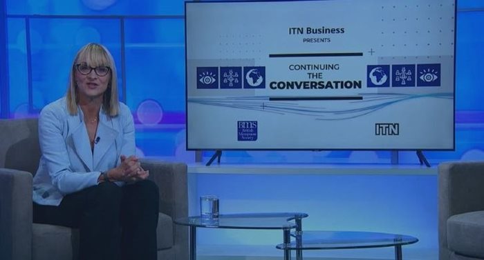 A woman in a light blue blazer sits on a chair in a studio setting. Beside her, a screen displays 