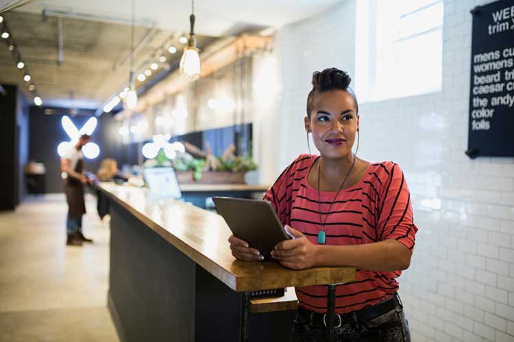 A person stands at a wooden counter holding a tablet, wearing headphones, in a modern, brightly lit space with hanging lights and decor