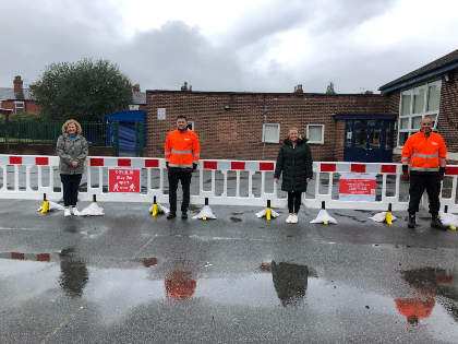Four people stand in a line, socially distanced, behind a barrier with red signage. Two wear orange safety jackets. The ground is wet and reflective.