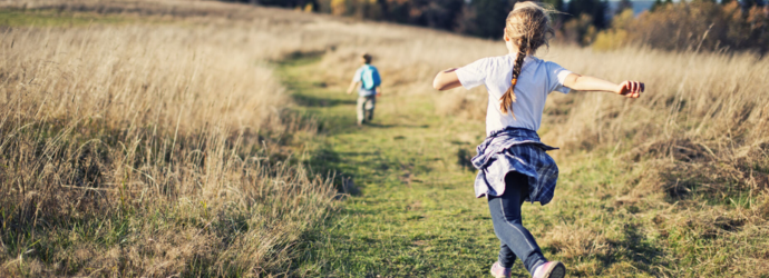 A girl and boy run seperately on a grassy path, surrounded by fields and trees in the background.