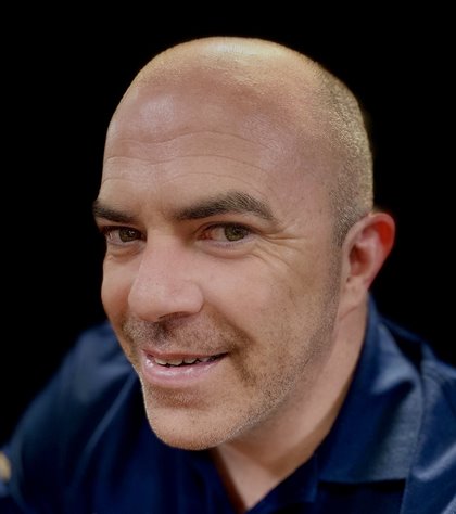 A bald man with a slight smile and stubble looks at the camera, wearing a dark shirt. The background is black, emphasizing his friendly expression.