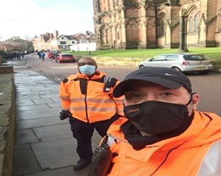 Two people wearing orange safety jackets and black masks stand on a sidewalk beside parked cars. A historic building and clear sky are in the background.