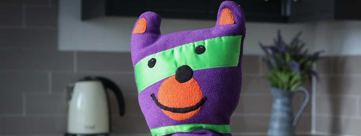 A close-up headshot of a Safety Seymour toy, infront of a kitchen setting backdrop