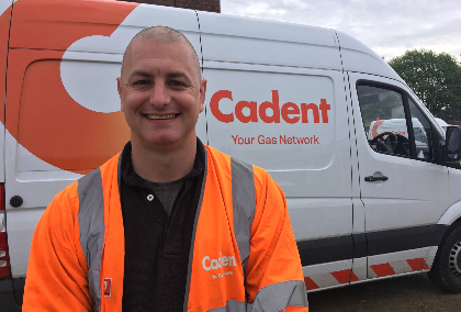 A smiling man in a high-visibility orange jacket stands in front of a white van with 