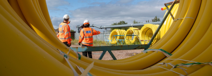 A person wearing a headset is smiling while looking at a computer screen in an office setting. They have long dark hair and are wearing a striped shirt. There are blurred figures and bright windows in the background.