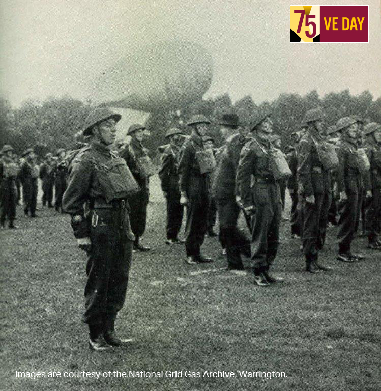 Group of World War II soldiers in uniform stand at attention in a field, with a large barrage balloon in the background. Tone is historical and solemn.