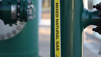 Close-up of a green industrial pipe labeled “Mixed Natural Gas” with bolts and fittings visible in the background.