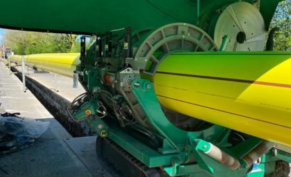 Equipment laying a large yellow pipeline in a trench under a green canopy. The scene is industrial and mechanical, with a clear blue sky visible.