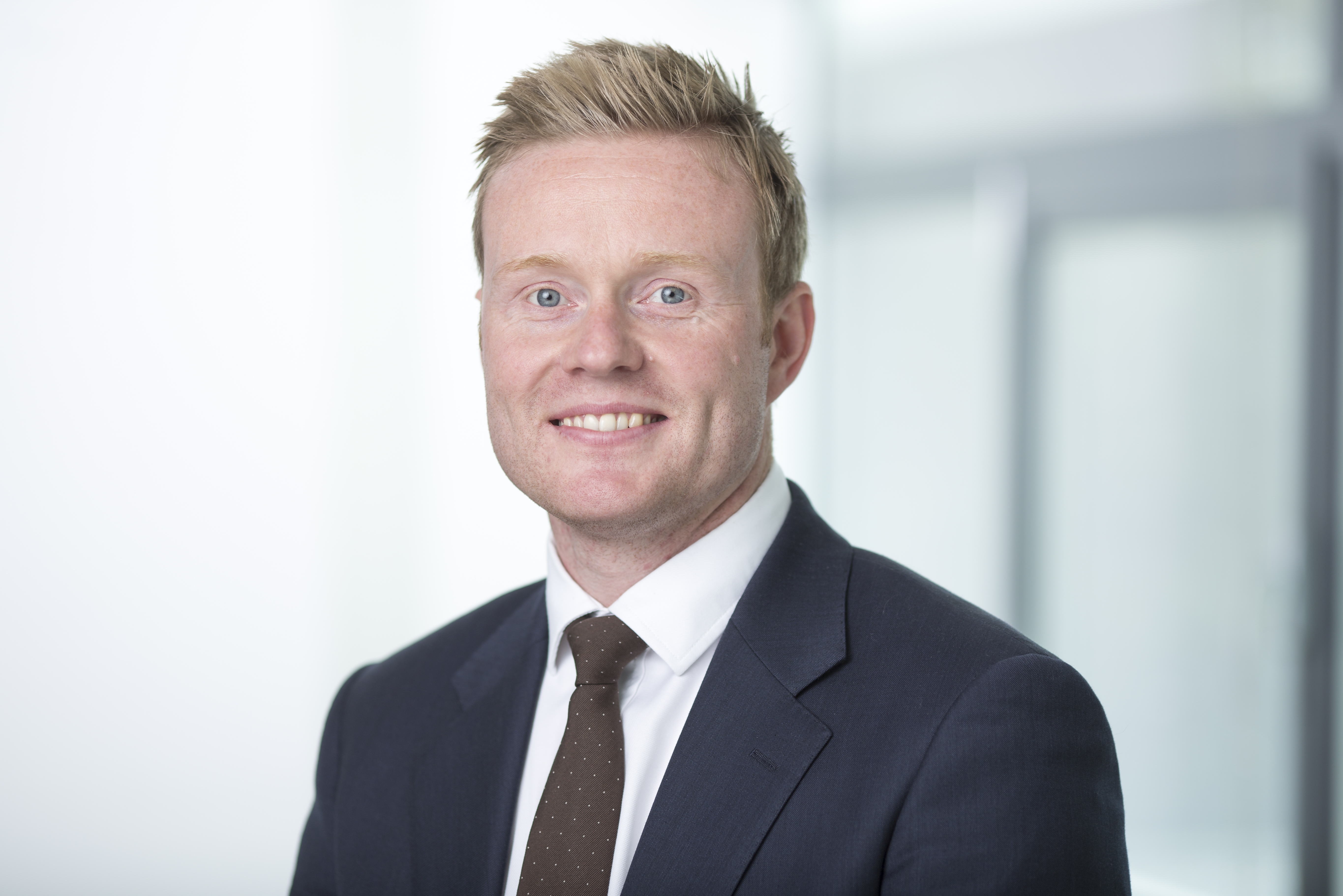 A man with light hair in a dark suit and brown tie smiles warmly. The background is softly blurred, suggesting a professional office setting.