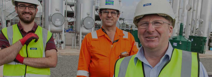 Workers wearing safety vests and protective gear stand in front of industrial equipment and piping at an outdoor facility.